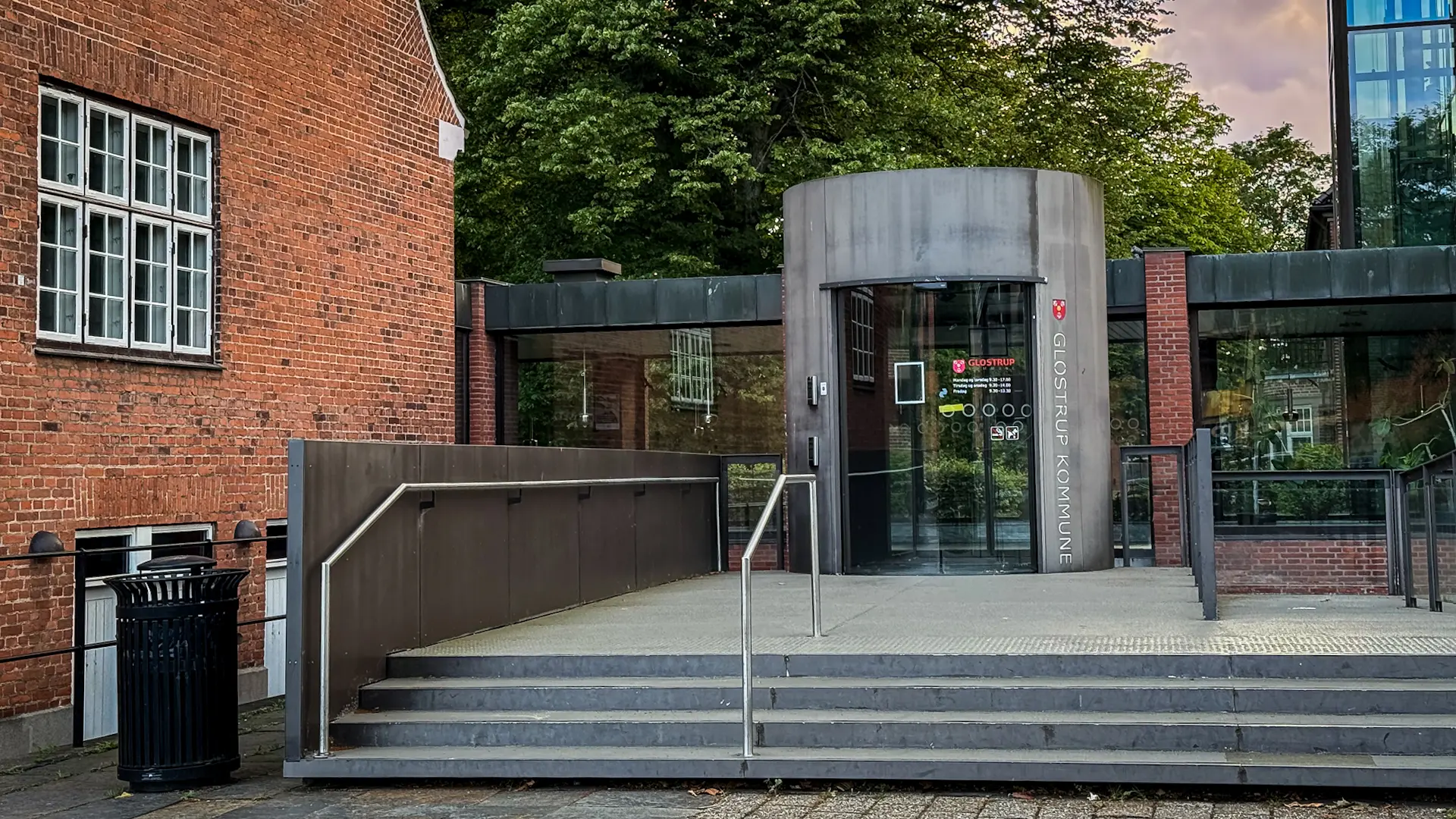 Grønt parkområde i Glostrup med træer, græsplæne og legeplads omgivet af boligblokke i baggrunden, der illustrerer kontrasten mellem byens grønne åndehuller og tæt bebyggelse.